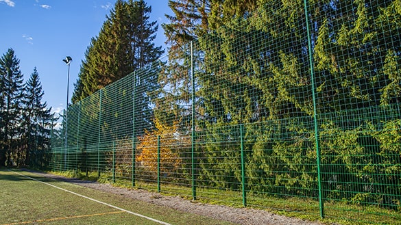 Vihreä elementtiaita palloilukentän ympärillä ja sen päälle asennettua korkeaa maalintaustaverkkoa estämässä palloja karkaamasta metsään.