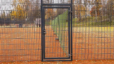 Musta elementtiaitaus ympäröi tenniskenttää, jonne pääsee mustan, abloy-lukittavan käyntiportin kautta.